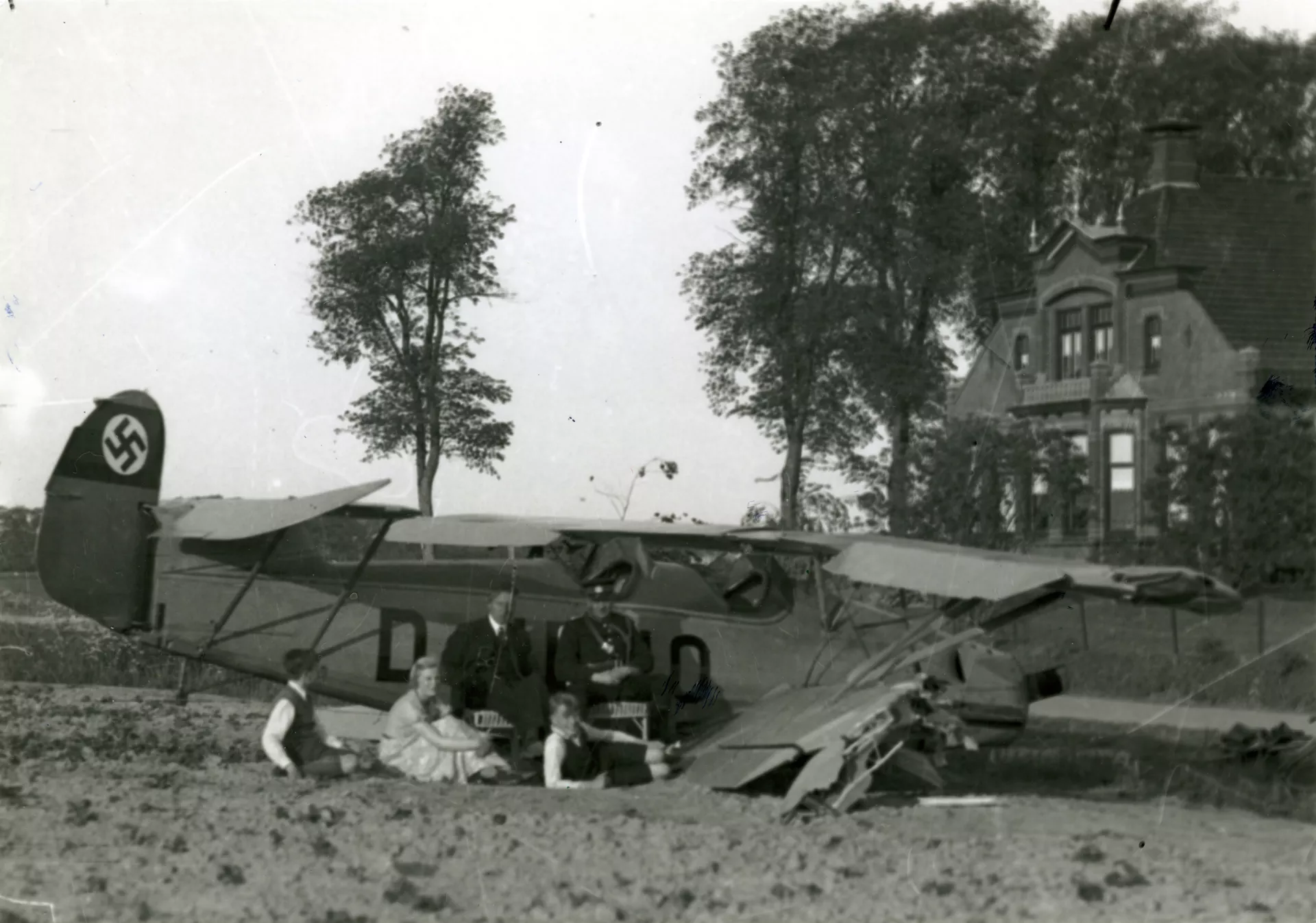 Een beschadigd Duits vliegtuig staat in een wei na een noodlanding. Een groep mensen zit naast het vliegtuig.