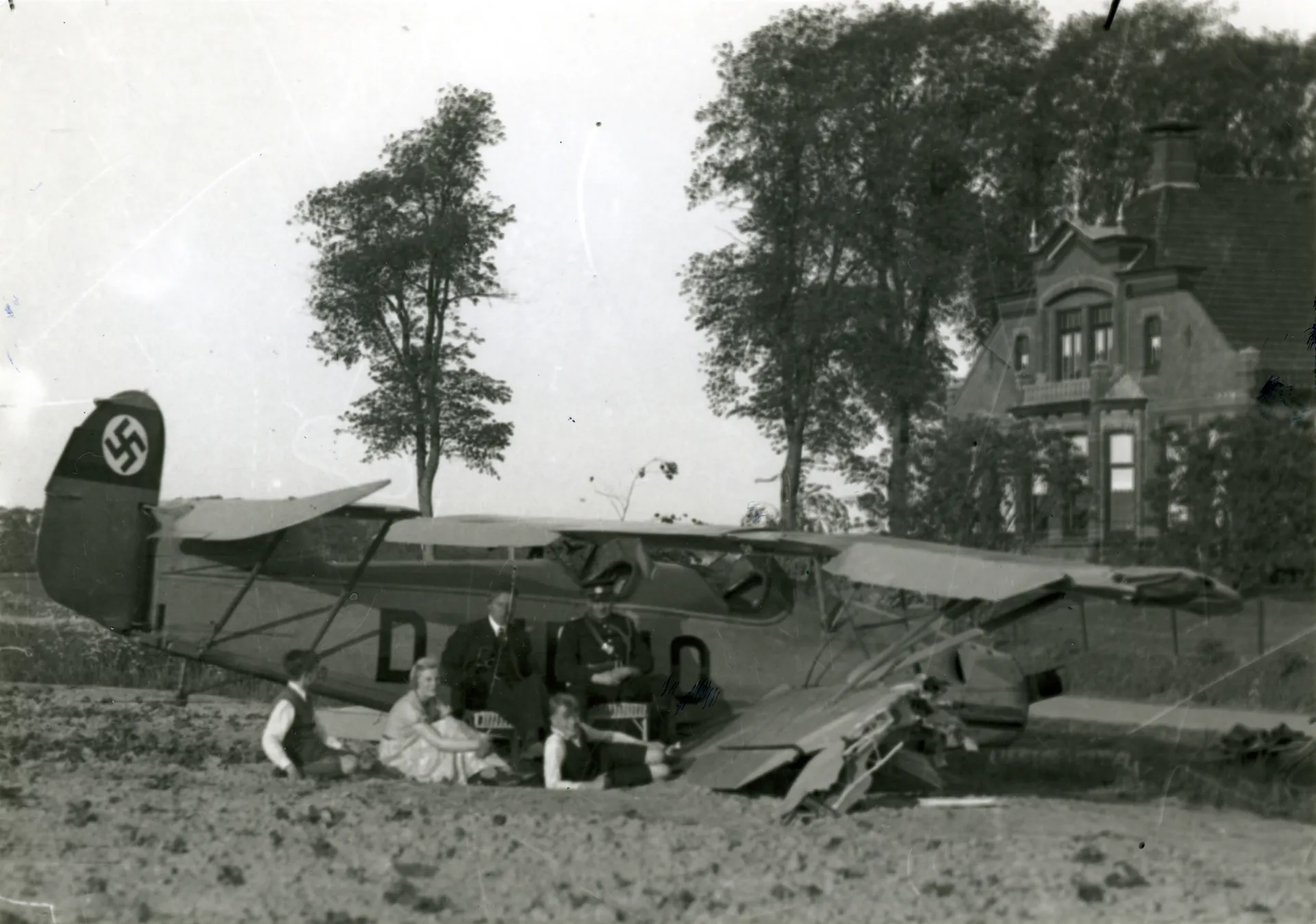 Een beschadigd Duits vliegtuig staat in een wei na een noodlanding. Een groep mensen zit naast het vliegtuig.