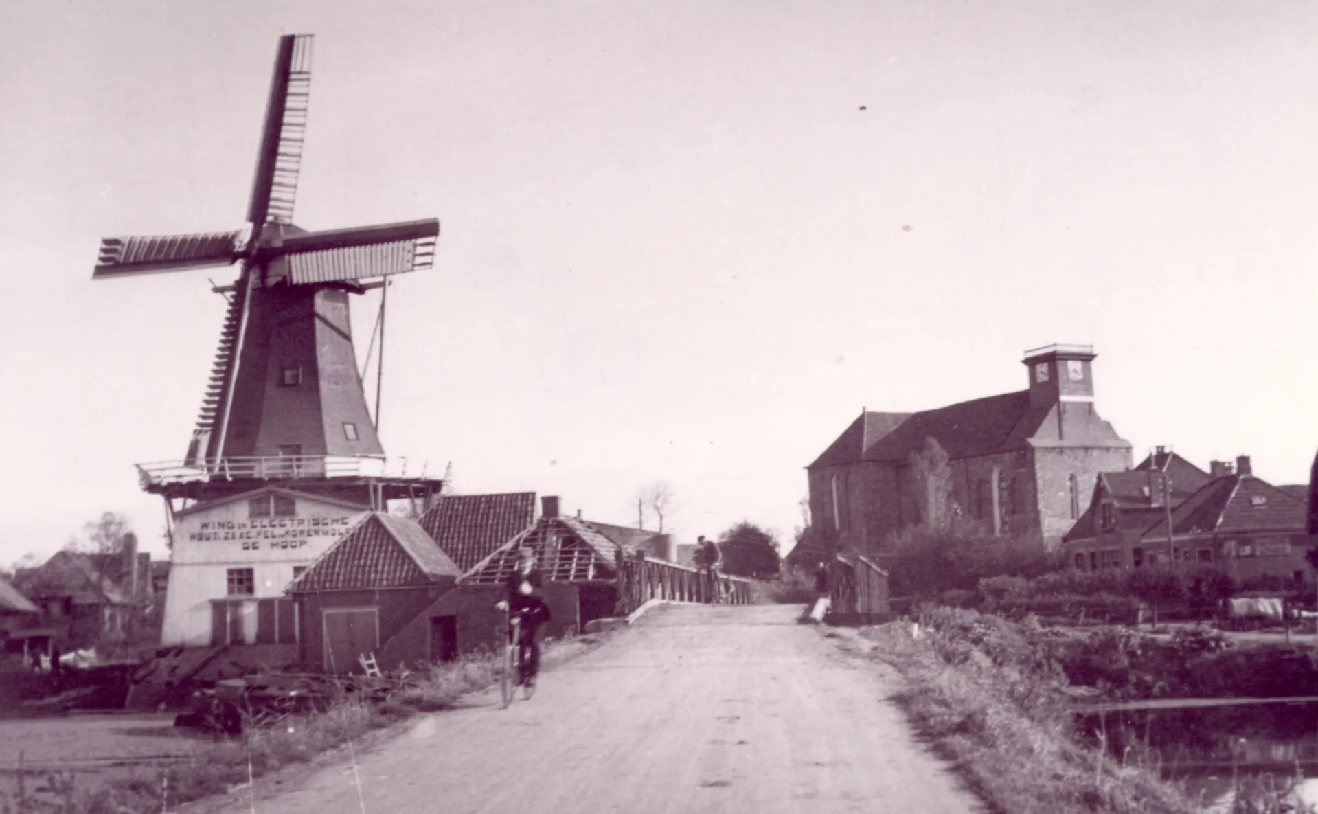 Een molen op de voorgrond met een kerk zonder toren op de achtergrond en huizen met beschadigde daken.. Een fietser fiets op de weg voor een noodbrug.