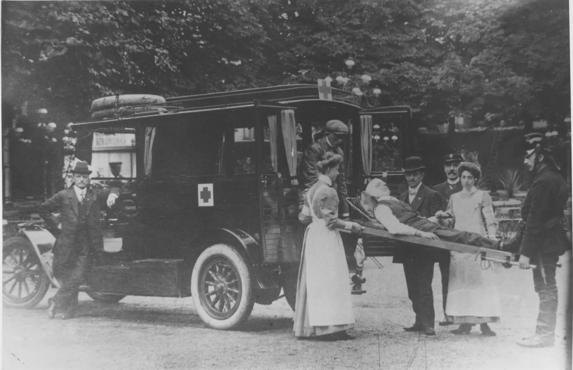 Zwart-wit foto van een ambulance van de ENNAM met ambulancepersoneel en een persoon op een brancard (1925).