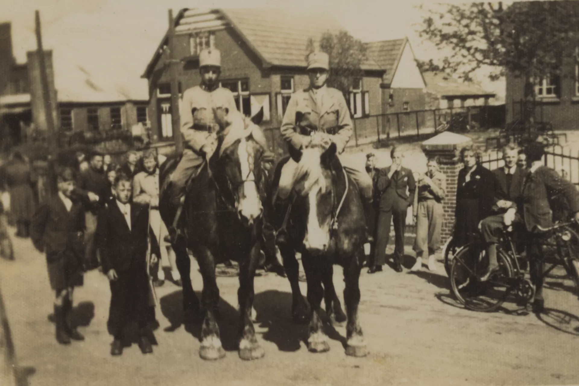Twee soldaten te paard omringd door mensen in een dorpsstraat.