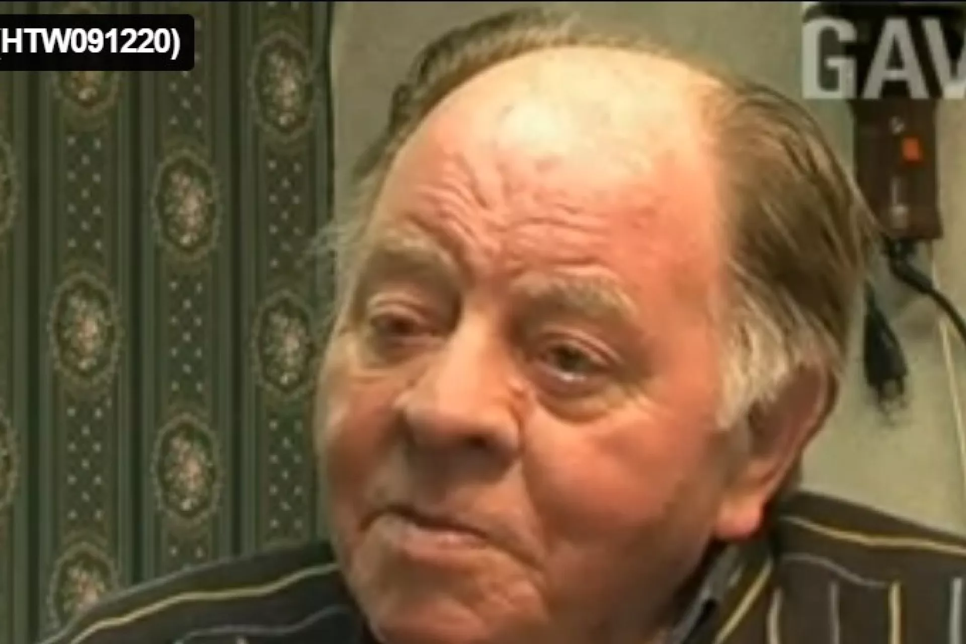 Een oudere man met een gestreepte blouse in een interieur met groen behang.