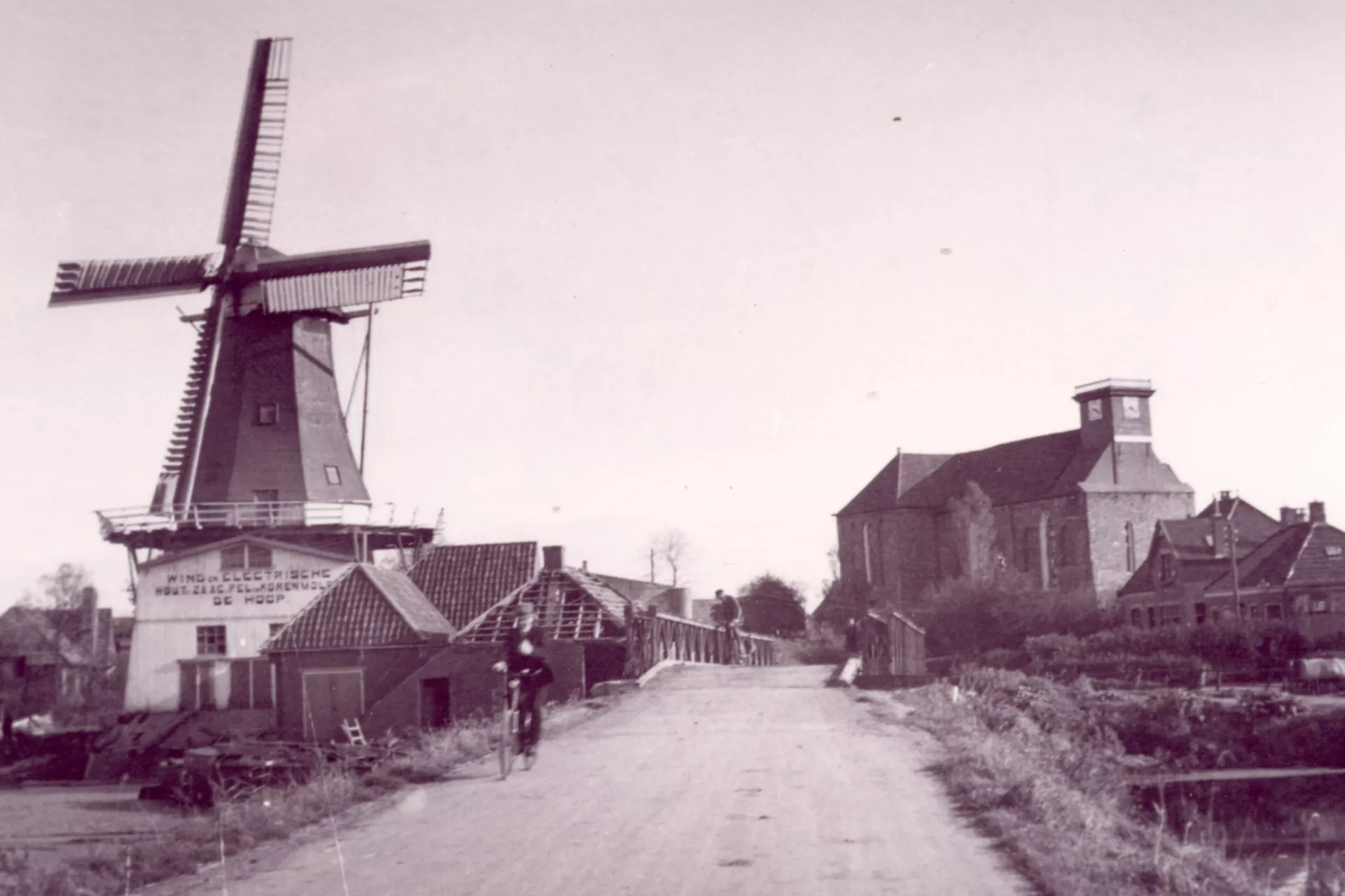 Een molen op de voorgrond met een kerk zonder toren op de achtergrond en huizen met beschadigde daken.. Een fietser fiets op de weg voor een noodbrug.